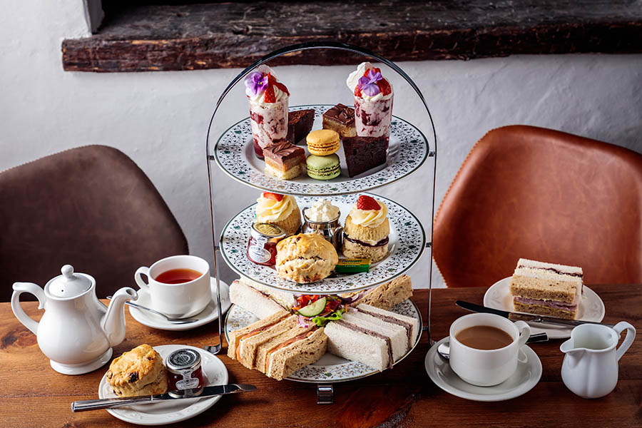 An elegant afternoon tea display on a three-tiered stand. The top tier is adorned with an array of cakes, including millionaire's shortbread and chocolate brownie. The middle tier features scones accompanied by small pots of jam and cream. The bottom tier holds an assortment of finger sandwiches.