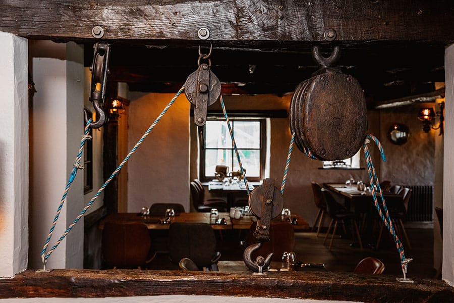 A view of the Crooklands Inn, featuring its charming interior with exposed wooden beams and vintage decorative pulley ropes in the foreground. The rustic and inviting ambiance of the inn is evident.